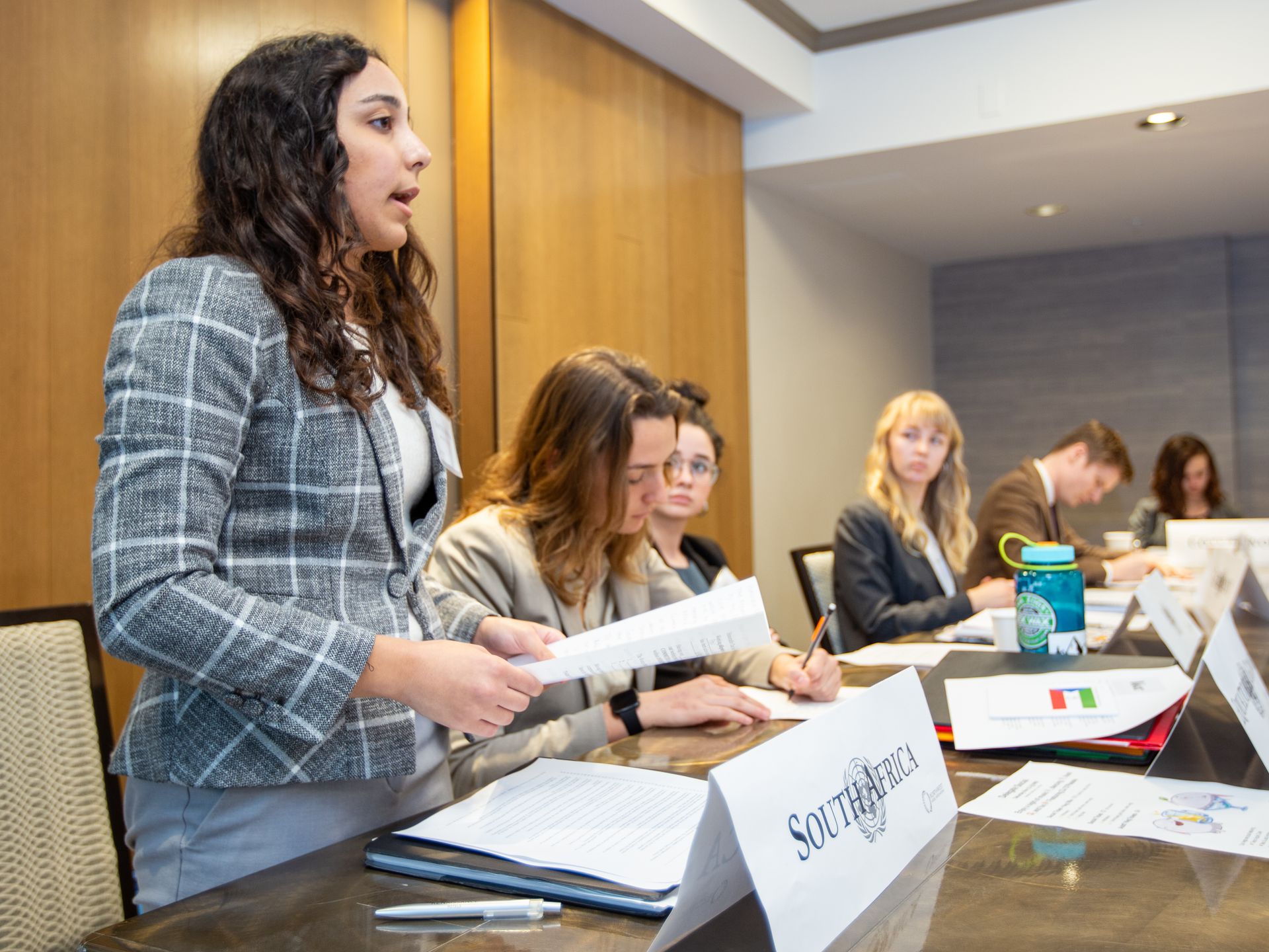A delegate speaking while fellow delegates take notes in committee